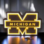 Mèche lumineuse LED en forme de logo Michigan avec inscription MICHIGAN en lettres jaunes sur fond noir, décoration intérieure lumineuse colorée, création 3D personnalisée.