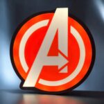 AVANT-PROPOS>A voir une lampe LED imprimée en 3D représentant le logo des Avengers d'Infinity 3D Print, illuminée en rouge et blanc, créant un effet visuel attrayant et personnalisé.