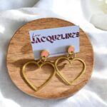 Épingles d'oreilles en forme de cœurs dorés avec détails 3D, présentées sur une carte avec le nom "Les Jacquelines", exposées sur un support en bois avec fond en tissu blanc.
