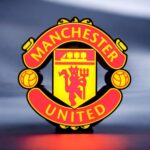 Logo LED lumineux du club de football Manchester United illuminé dans l'obscurité. La lampe 3D met en valeur le symbole emblématique du club avec ses couleurs rouges et jaunes et ses détails précis.